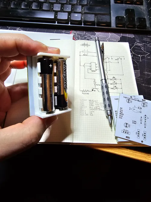 Hand-drawn circuit schematic in a notebook with a 3D-printed 2xAA battery holder in the foreground