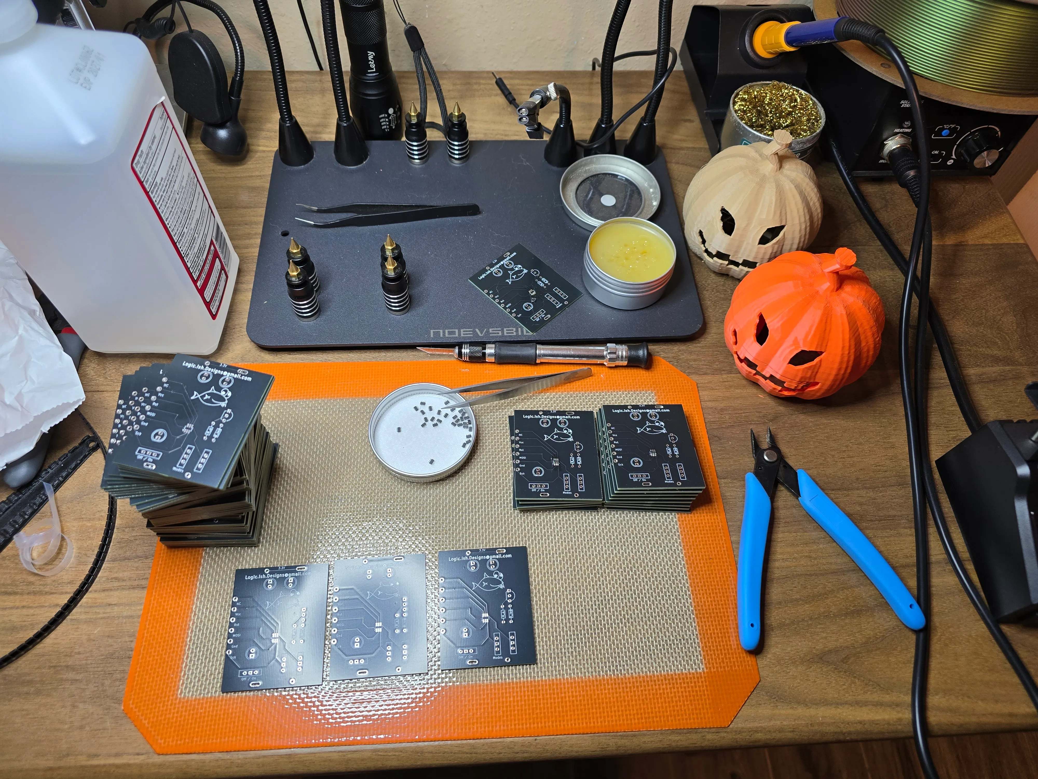 Workbench with dozens of finished custom PCBs and two 3D-printed jack-o’-lantern shells